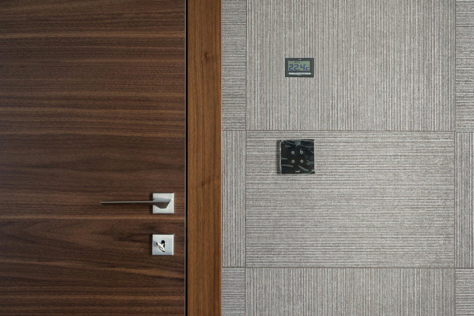 A digital thermostat displaying 22.4°C mounted on a textured gray wall next to a modern wooden door with a metal handle and lock.