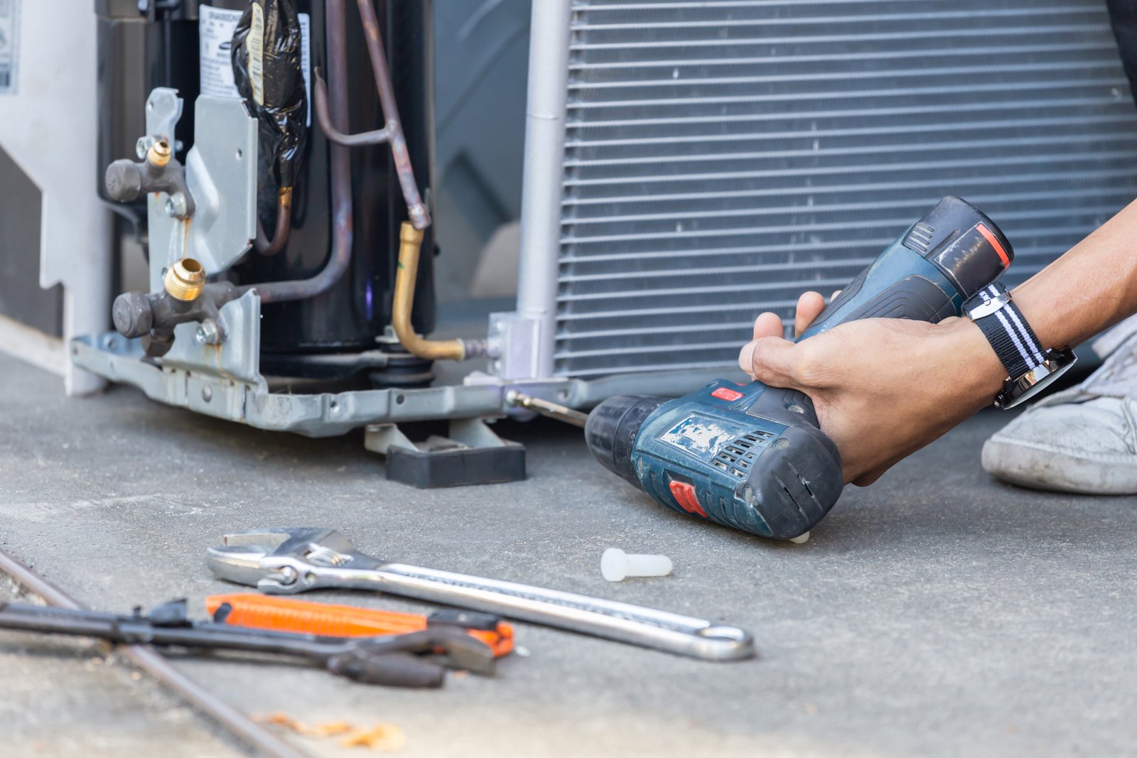 HVAC repair technician using a power drill to fix an air conditioning compressor unit, with tools scattered nearby for repair work.