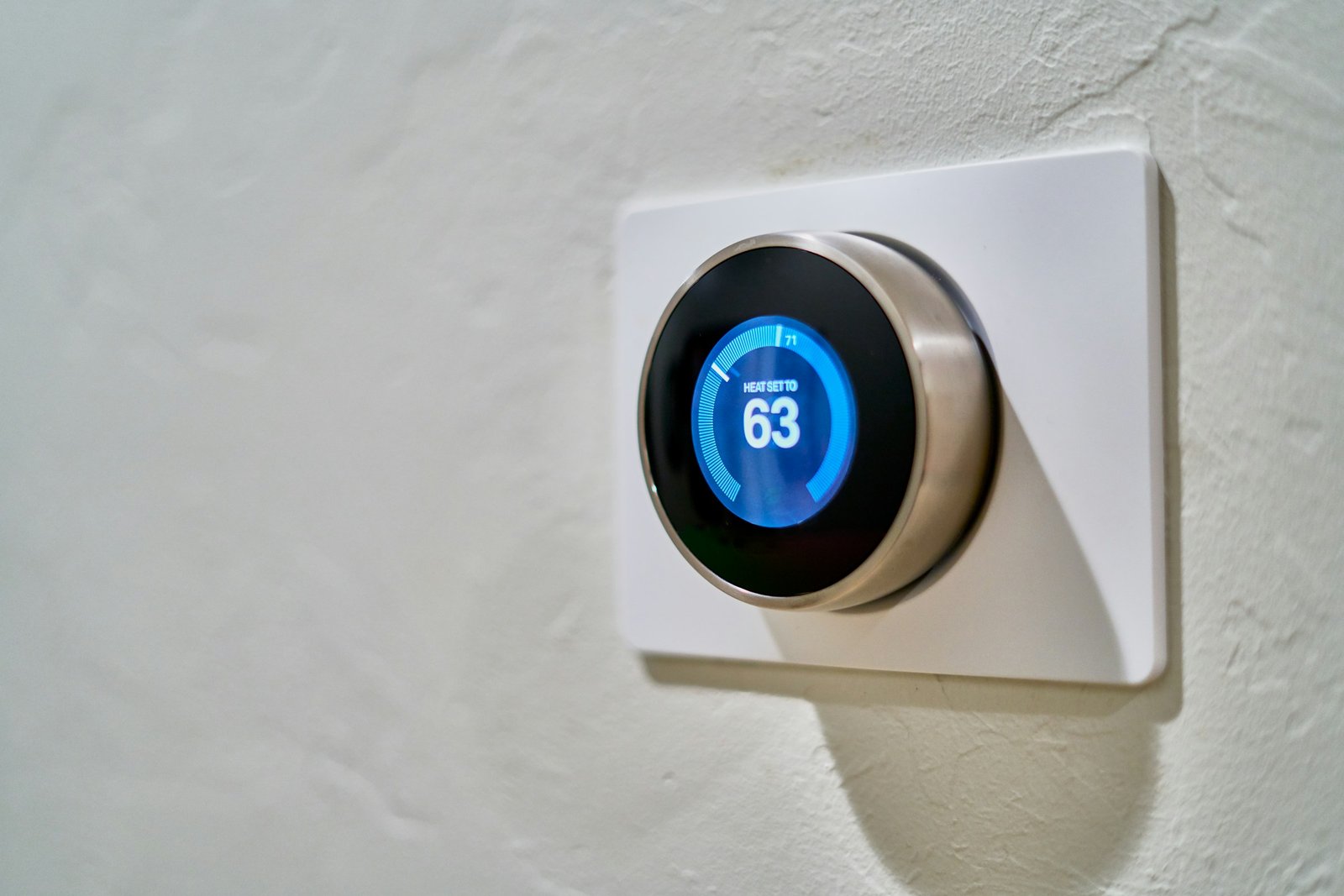 A modern smart thermostat mounted on a white textured wall, displaying a heating setting of 63 degrees Fahrenheit.