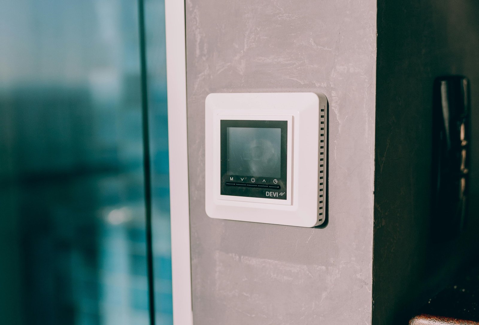 A wall-mounted digital thermostat installed on a gray concrete wall near a glass door, reflecting natural light.
