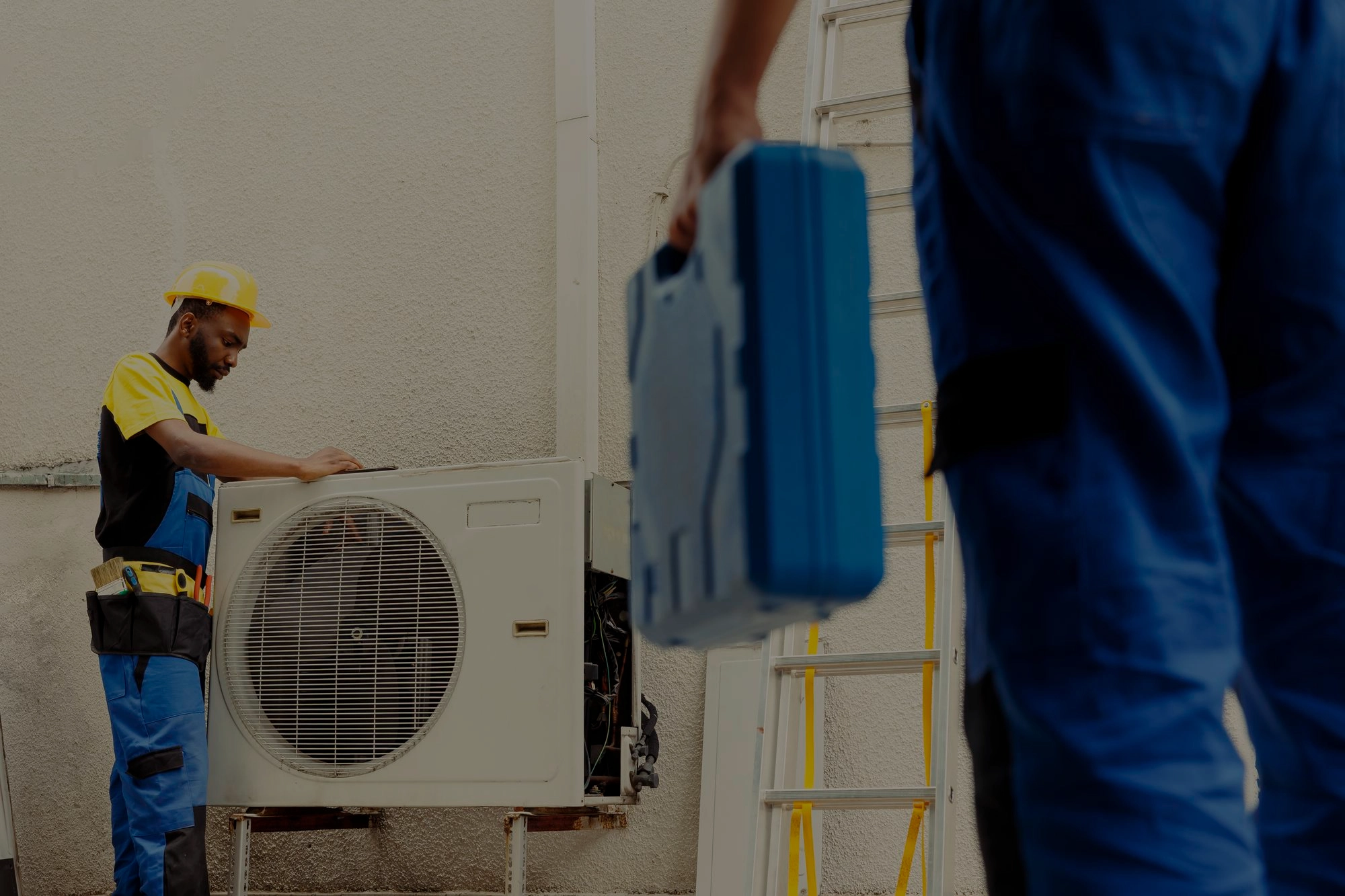 HVAC technicians in safety gear working on an outdoor air conditioning unit, with one adjusting the system and the other holding a tool case